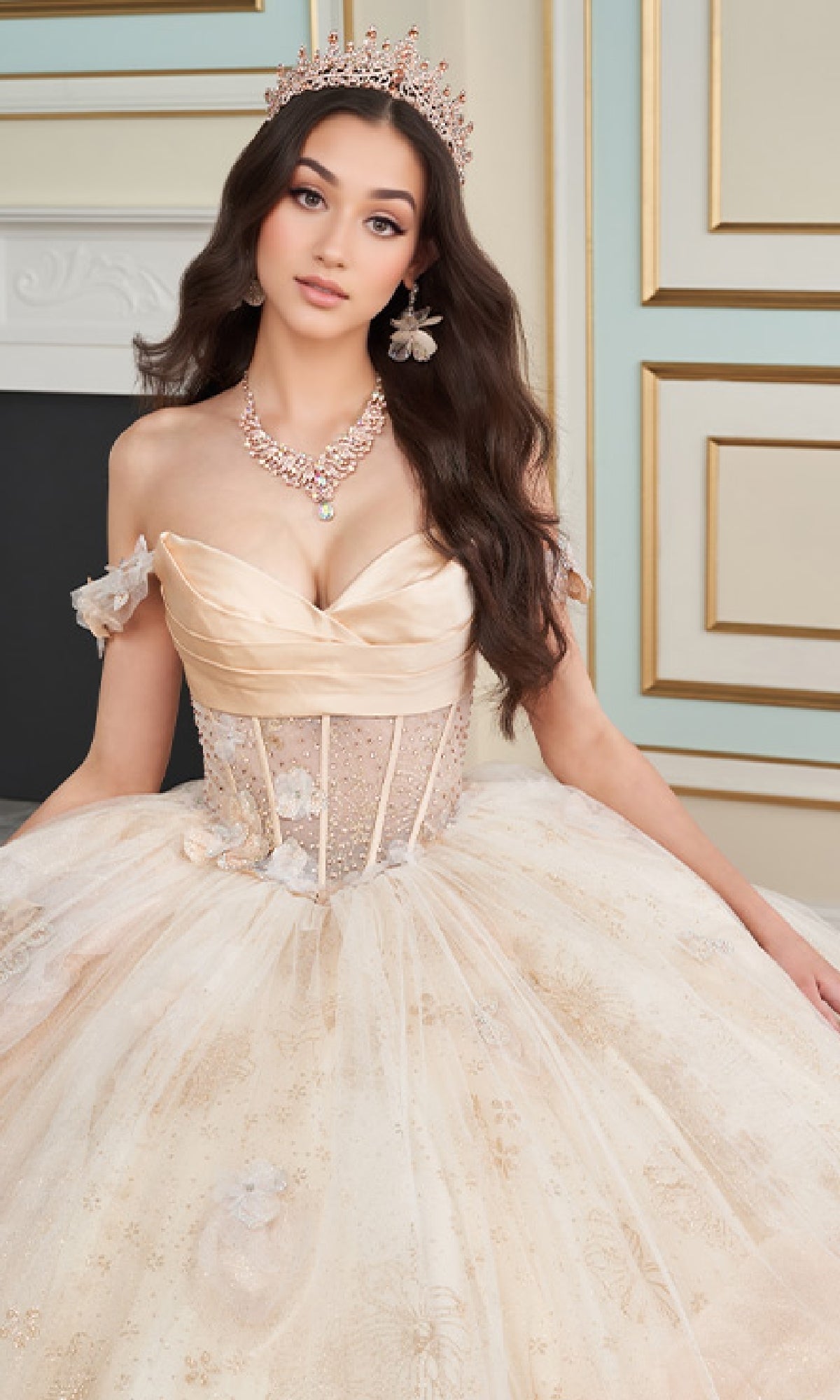 A woman wearing the Quinceanera Dress PR30159 By Princesa, featuring a cream color and corset bodice, poses indoors with a jeweled crown and necklace against an ornate background.